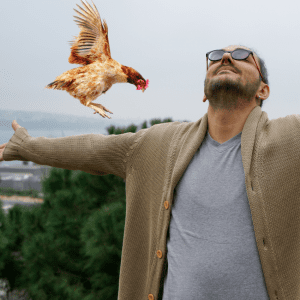 A man with sunglasses and a cardigan joyfully spreading his arms as a chicken flies is about to land on his arm.