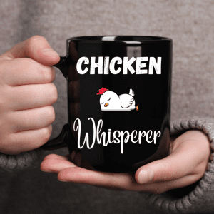 The image shows a person holding a black mug that has the words "CHICKEN Whisperer" printed on it in white stylized text. Accompanying the text is an adorable graphic of a white chicken with a red comb, looking content. This mug has a fun, playful feel and would likely appeal to chicken enthusiasts or owners who have a good sense of humor about their bond with their poultry.