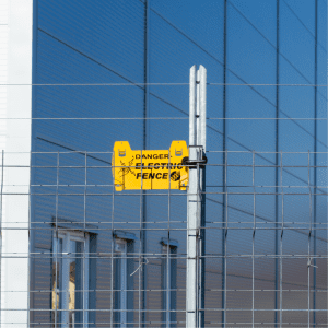 The image depicts a bright yellow warning sign with the words "DANGER ELECTRIC FENCE" and symbols indicating the risk of shock. The sign is mounted on a metal post that's part of a tall, multi-wired electric fence. The background shows a modern building with a blue facade, suggesting that the electric fence is part of a contemporary, secure establishment, possibly to protect property or livestock.