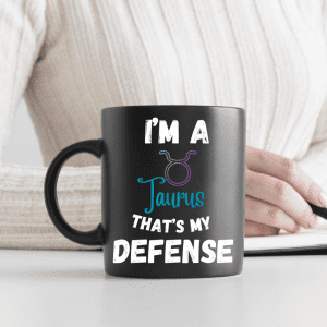 "A black mug with the phrase 'I'm a Taurus, that's my defense' and the Taurus astrological symbol, held by a person."