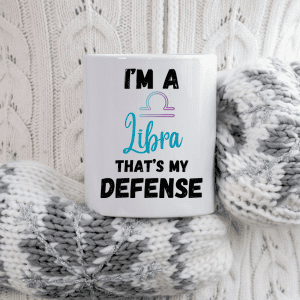 Horoscope mugs - the image displays a white mug with the text "I'M A Libra THAT'S MY DEFENSE" printed on it. Above the word "Libra," there is an icon representing the Libra zodiac sign, featuring the traditional scales in a gradient of pastel colors. The mug is nestled between what appears to be a pair of cozy knitted winter gloves with snowflake decorations, against a cable-knit sweater background, giving off a warm, comfortable vibe. This mug would likely appeal to those who enjoy expressing their zodiac pride, especially Libras, and it fits nicely into a cozy, seasonal setting.