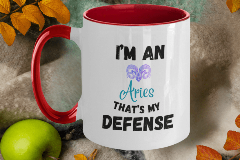 The image shows a white mug with a red interior and handle, with the text "I'M AN Aries THAT'S MY DEFENSE" printed on it. Above the word "Aries," there's an illustration of the Aries zodiac sign symbol in a colorful design. The mug is set against a background that includes a green apple and autumn leaves, suggesting a cozy, seasonal atmosphere. This type of mug could be a personalized item, part of a zodiac-themed collection, and might appeal to those who identify with their astrological sign, particularly Aries in this case.