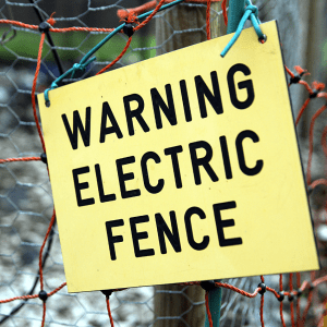 The image shows a warning sign with bold black lettering on a yellow background that reads "WARNING ELECTRIC FENCE." The sign is attached to a fence with red and blue knots, indicating that the fence is electrified. This serves as a caution to people to keep a safe distance and not to touch the fence, which is designed to prevent animals or unauthorized persons from crossing the boundary.