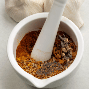 : A mortar and pestle with a mixture of various spices, including cloves, being ground. Garlic bulbs are visible in the background.